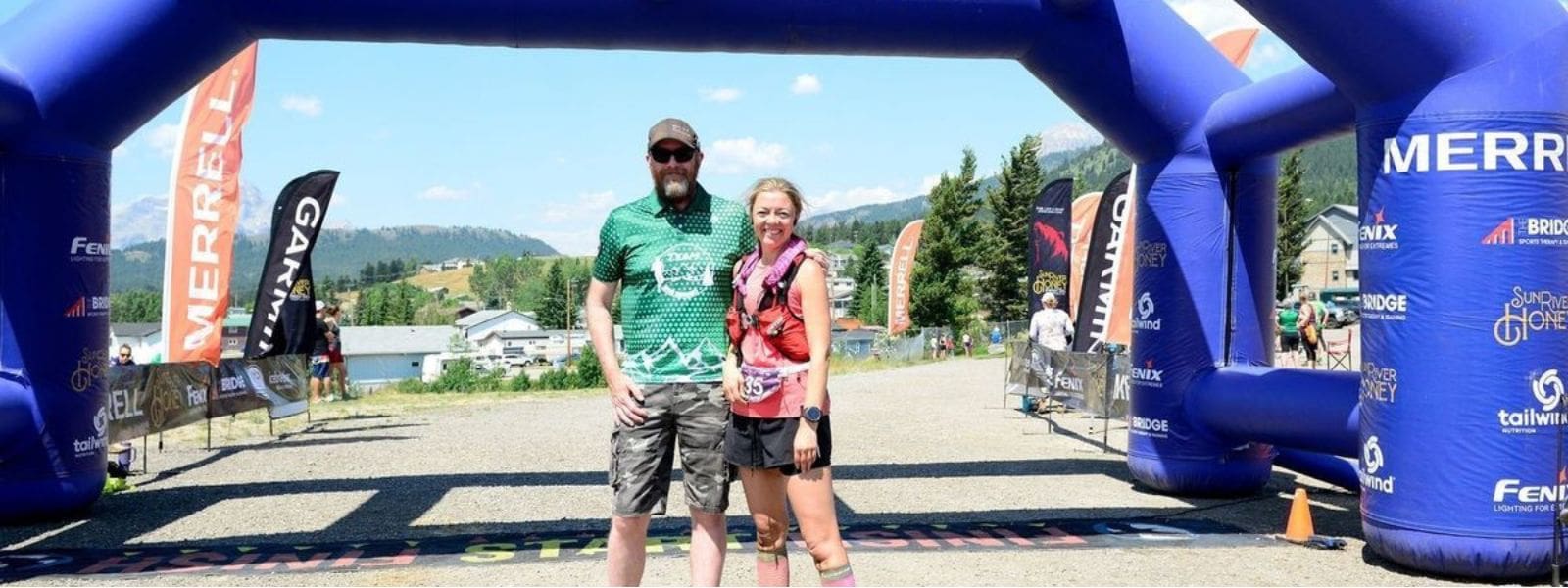 Coach JW and an ultra trail athlete standing together under an inflatable arch at a trail race finish line with mountains behind.